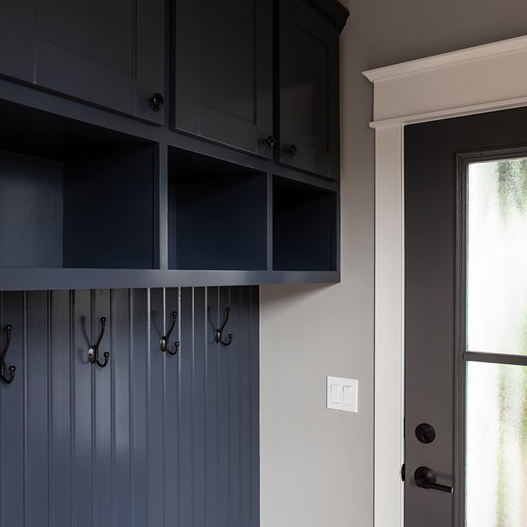 Navy blue cabinetry with coat hooks and gray walls in this beautiful mudroom by BDS Design Build Remodel.