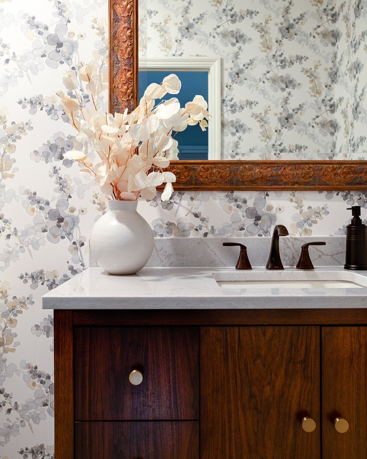 Small bathroom with wallpaper, mirror, wooden vanity and a flower vase on top of the white counter top.