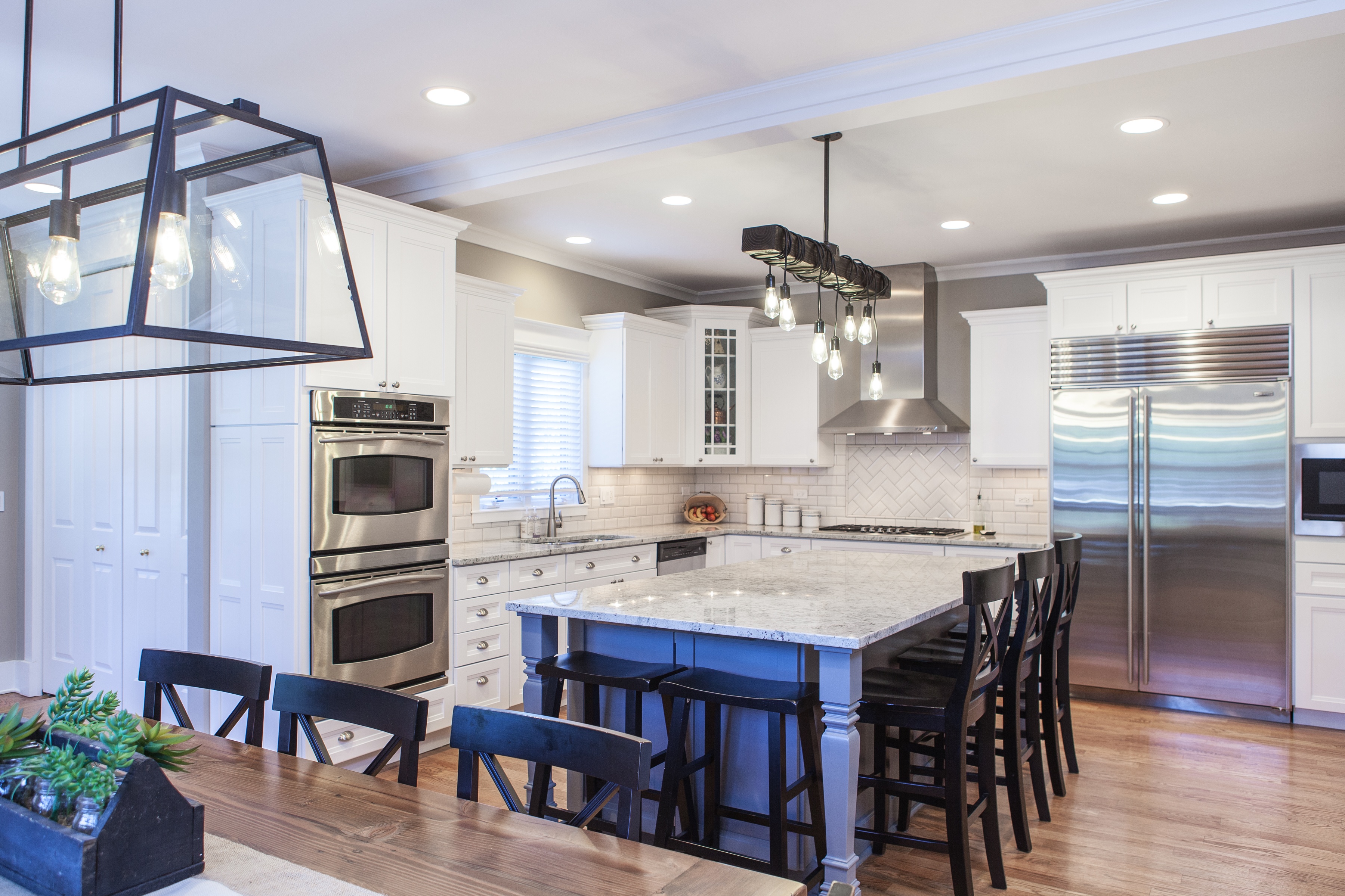 farmhouse kitchen with white upper cabinets and a slate blue island. Black chairs and hardwood floors.