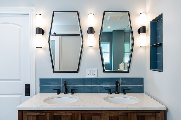 Primary bathroom with dual, black-framed geometric mirrors, up-down wall sconces, blue subway tile, black bathroom hardware and a custom wood vanity.